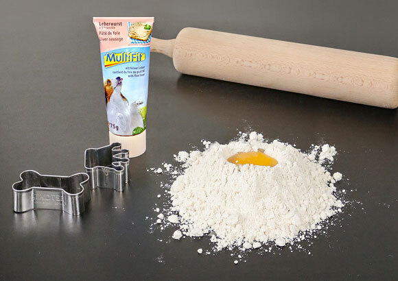 Backzutaten für Hundekekse mit Mehl, Ei, Leberwurstpaste, Ausstechformen in Hunde- und Knochenform sowie Nudelholz auf dunklem Untergrund