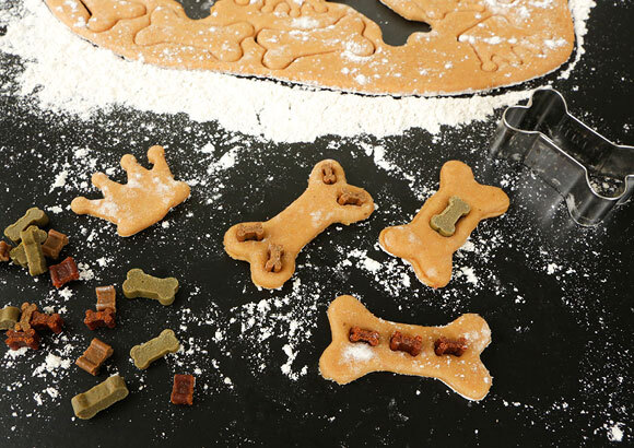 Selbstgemachte Hundekekse in Knochenform mit Leckerlis, Ausstechform und Mehl auf schwarzer Arbeitsfläche