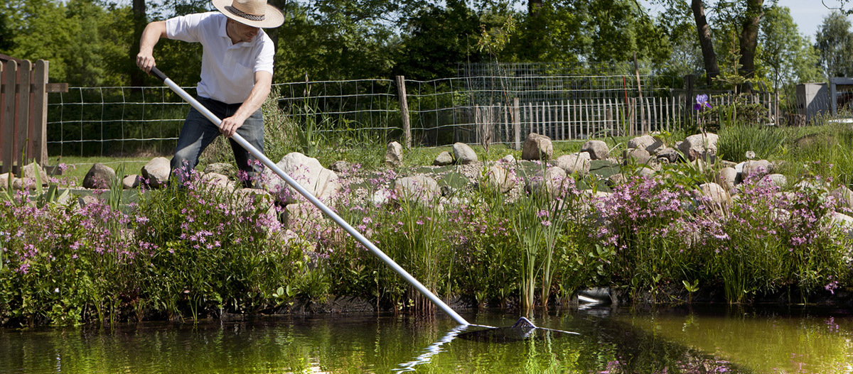 Ein Mann holt mit einem Kescher etwas aus dem Wasser. Mann mit Strohhut reinigt Teich im Garten mit langem Kescher, umgeben von blühenden Pflanzen und Steinen
