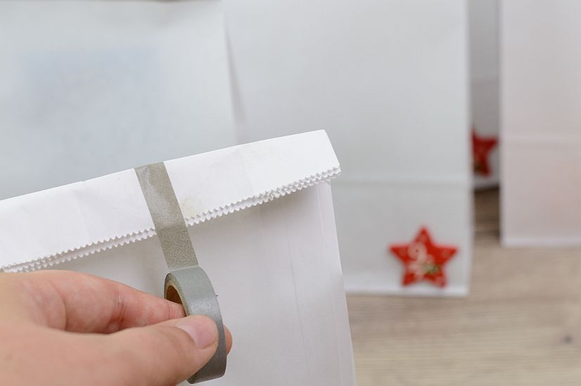 Hand klebt silbernes Klebeband auf weiße Papiertüte mit rotem Stern im Hintergrund, Geschenkverpackung und Bastelidee