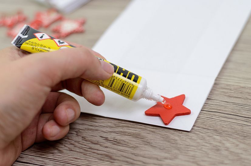 Hand trägt Klebstoff auf einen roten Papierstern auf, Bastelprojekt auf Holztisch