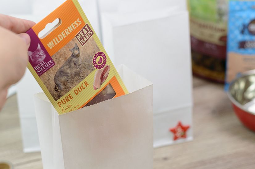 Hand legt Real Nature Wilderness Meat Flakes Pure Duck Katzenleckerlis in eine weiße Papiertüte