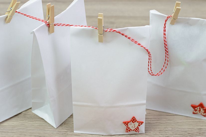 Weiße Papiertüten mit roten Sternen und Zahlen, aufgehängt an einer rot-weißen Schnur mit Holzklammern, Adventskalender-Dekoration.