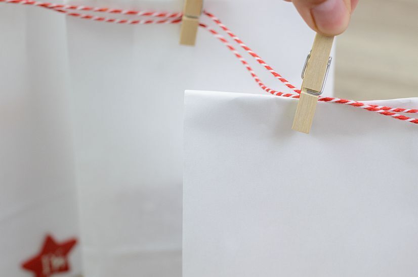 Hand befestigt weiße Papiertüte mit Holzklammer an rot-weißem Schnur für Adventskalender oder Geschenkverpackung