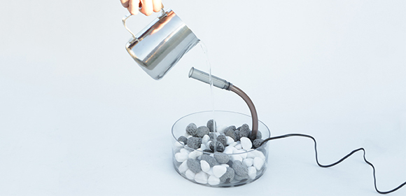 Moderner Luftbefeuchter mit schwarzen und weißen Steinen in einem transparenten Behälter, Wasser wird aus einem Metallkrug gegossen, minimalistisches Design.