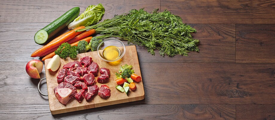 Frisches rohes Fleisch mit Karotten, Gurke, Apfel, Brokkoli und Olivenöl auf einem Holzbrett auf einem dunklen Holztisch.