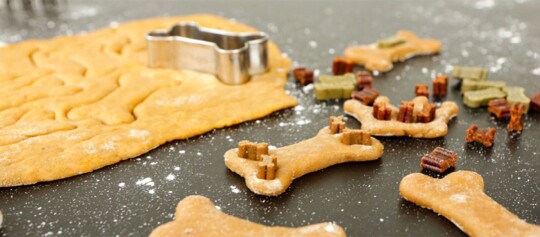 Hundeknochenförmige Kekse aus Teig mit Metall-Ausstecher und bunten Hundeleckerlis auf dunkler, bemehlter Arbeitsfläche