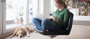 Junge Frau sitzt auf einem modernen Stuhl und tätigt Online-Einkäufe mit Kreditkarte und Laptop, während ihr entspannter Golden Retriever neben ihr liegt.