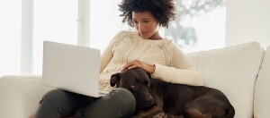 Junge Frau arbeitet entspannt mit Laptop auf dem Sofa und streichelt ihren schwarzen Hund