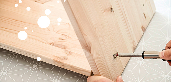 Anleitung DIY Katzenhaus Nahaufnahme einer Person, die mit einem Schraubendreher ein Möbelstück aus hellem Holz zusammenbaut, auf geometrisch gemustertem Untergrund