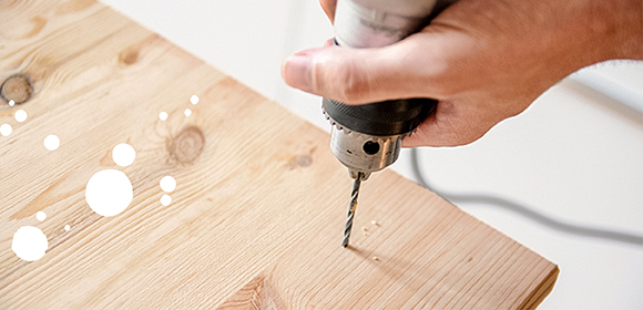 Anleitung DIY Katzenhaus Hand hält einen elektrischen Bohrer und bohrt ein Loch in eine helle Holzplatte mit sichtbarer Maserung.