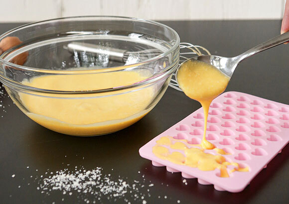 Anleitung um Snacks für Katzen selber zu backen Gelber Kuchenteig wird mit einem Löffel in eine rosa Silikonform mit kleinen Herzformen gegossen, neben einer Glasschüssel mit Teig und einem Schneebesen