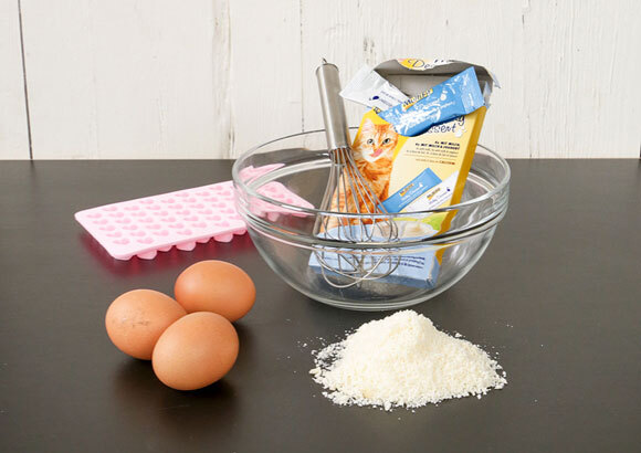 Anleitung um Snacks für Katzen selber zu backen Küchenutensilien mit drei Eiern, einer Glasschüssel mit Schneebesen und Katzenfutter, weißem Pulver und rosa Silikonform mit Herzmotiven.