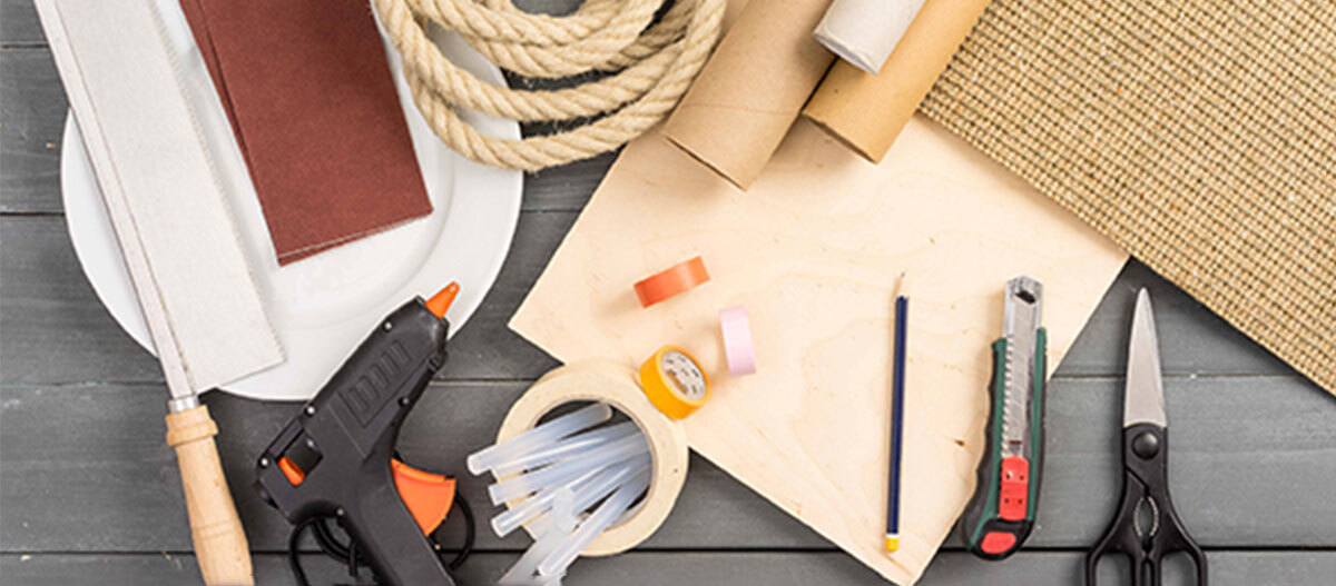 Anleitung DIY Spielzeug für Katzen Verschiedene Bastel- und Heimwerkerwerkzeuge wie Handsäge, Heißklebepistole, Klebebänder, Seil, Cutter, Schere und Bleistift auf einem Holztisch