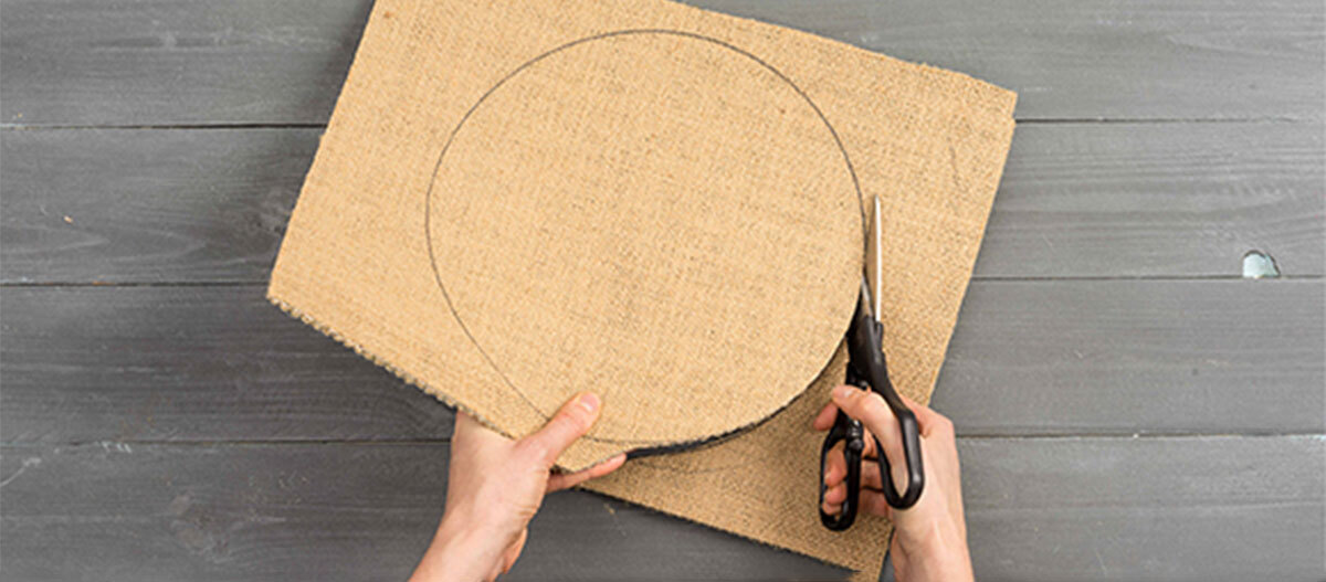 Anleitung DIY Spielzeug für Katzen Person schneidet einen Kreis aus Jutestoff mit einer Schere auf einem grauen Holztisch