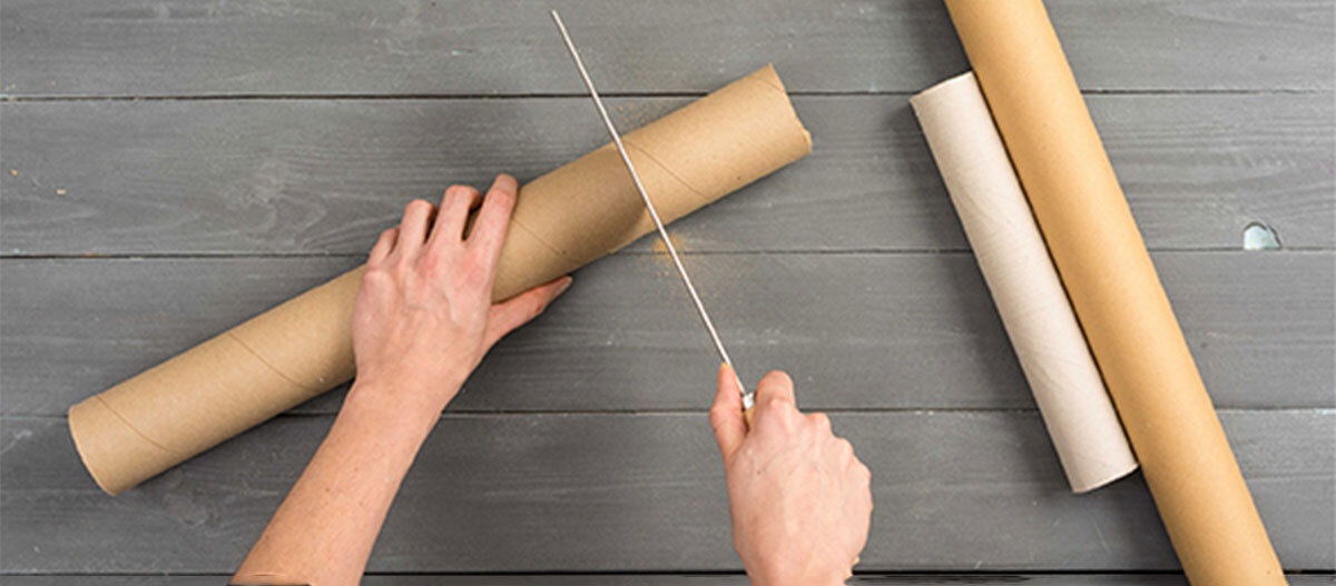 Anleitung DIY Spielzeug für Katzen Person schneidet große Papprolle mit Handsäge auf grauem Holztisch, zwei weitere Papprollen liegen daneben