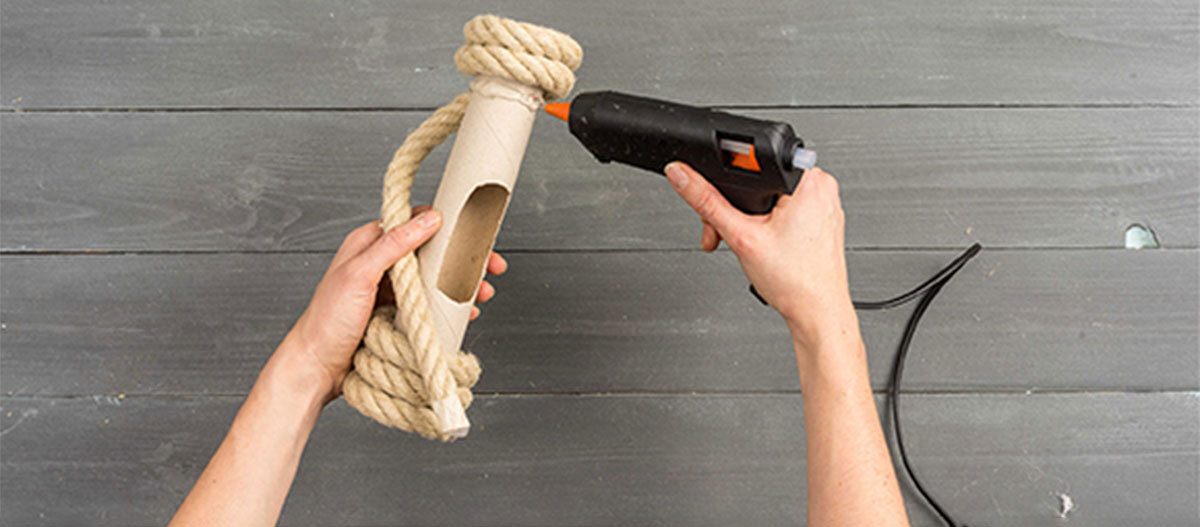 Anleitung DIY Spielzeug für Katzen Person klebt dickes Naturseil mit Heißklebepistole an Papprolle auf grauem Holztisch DIY Bastelprojekt