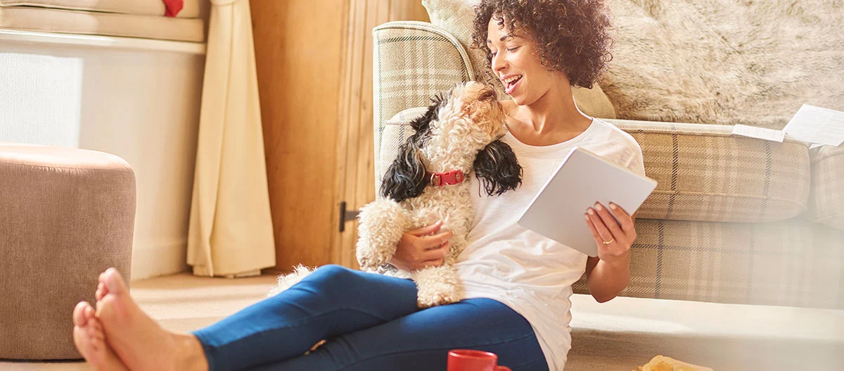 Junge Frau sitzt auf dem Boden und liest auf einem Tablet, während sie liebevoll mit ihrem kleinen schwarz-weißen Hund mit rotem Halsband kuschelt.