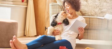 Junge Frau sitzt auf dem Boden und liest auf einem Tablet, während sie liebevoll mit ihrem kleinen schwarz-weißen Hund mit rotem Halsband kuschelt.