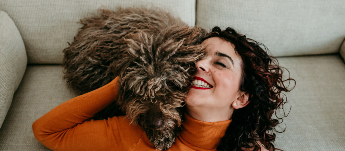 Glückliche Frau mit lockigem Haar kuschelt liebevoll mit großem, flauschigem braunem Hund auf einem hellen Sofa