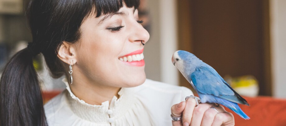 Junge Frau lächelt glücklich, während sie einen kleinen blau-weißen Papagei auf ihrer Hand hält.
