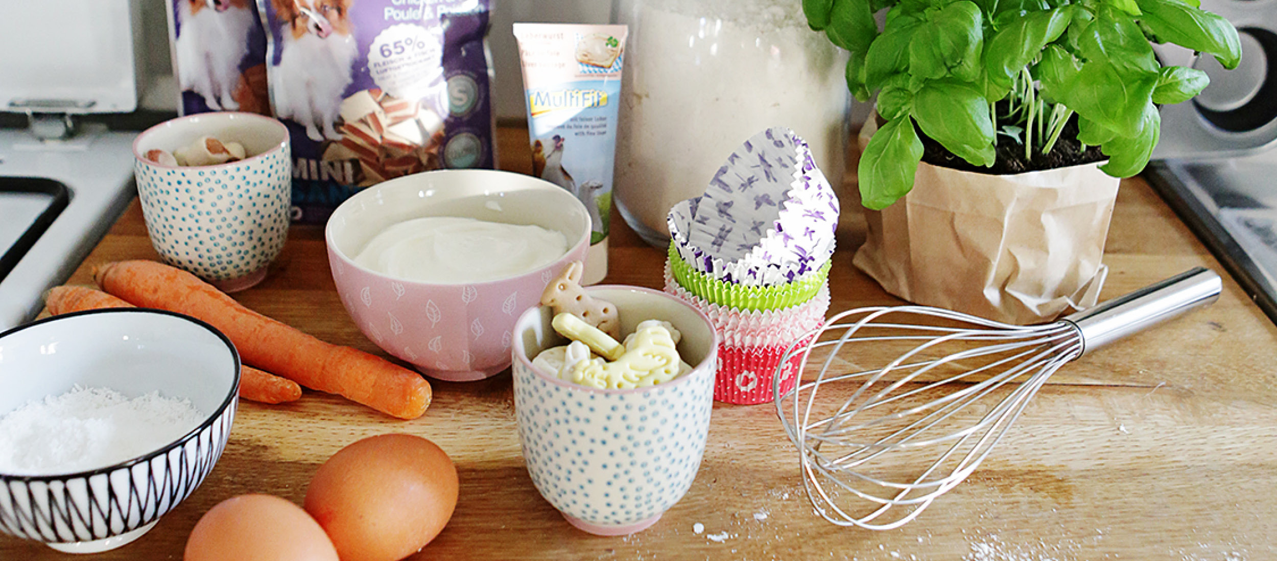 0220_Muffins_DIY_Online_1200x527px Backzutaten und Küchenutensilien auf Holzarbeitsplatte mit Karotten, Eiern, Mehl, Joghurt, Hundekeksen, Basilikum und Schneebesen