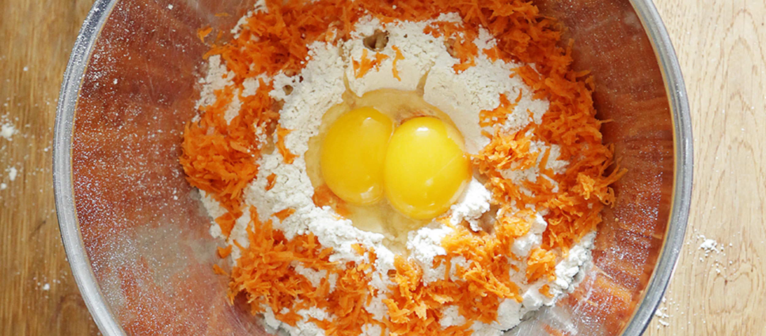 0220_Muffins_DIY_Online_1200x527px2 Backschüssel mit Mehl, geriebenen Karotten und zwei rohen Eiern für Karottenkuchen-Rezept