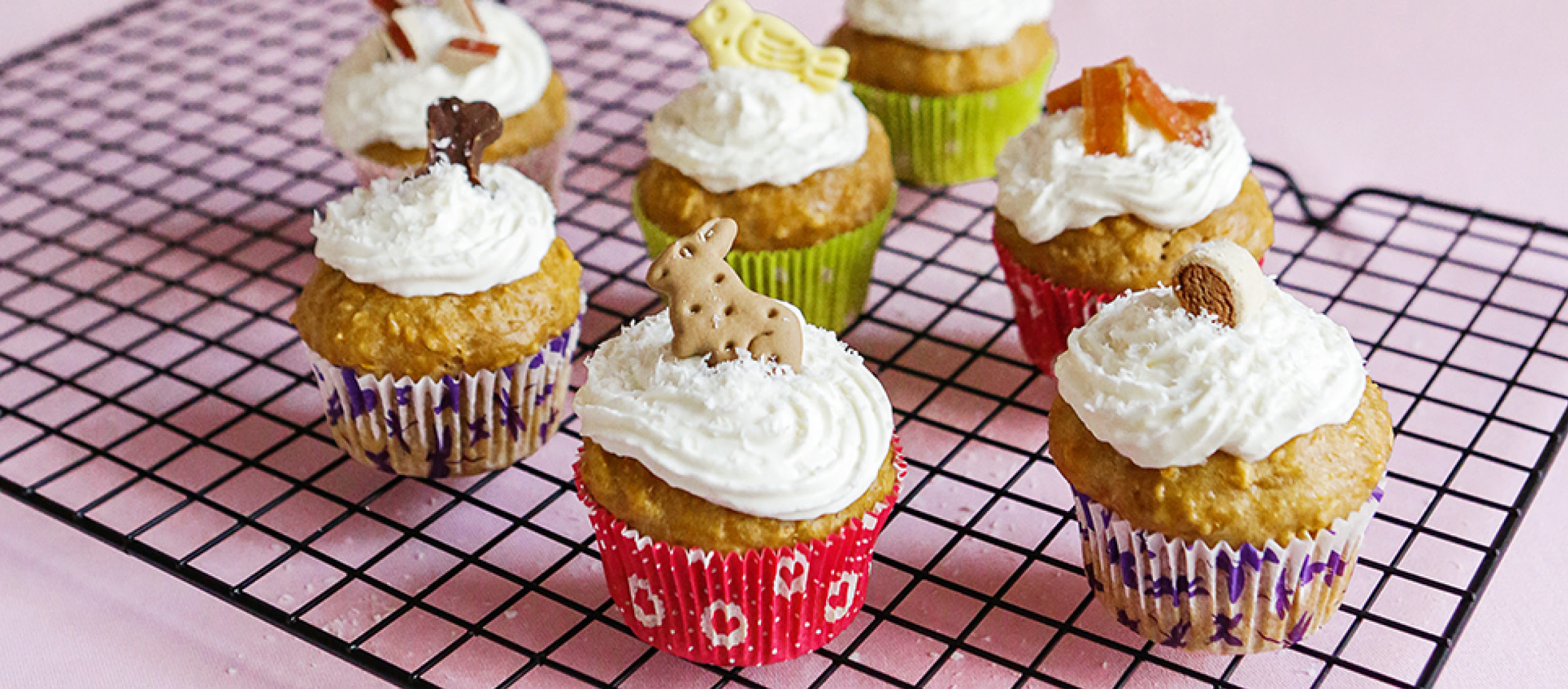 0220_Muffins_DIY_Online_1200x527px3 Bunte Cupcakes mit weißem Zuckerguss und verschiedenen essbaren Dekorationen auf einem schwarzen Abkühlgitter vor rosa Hintergrund