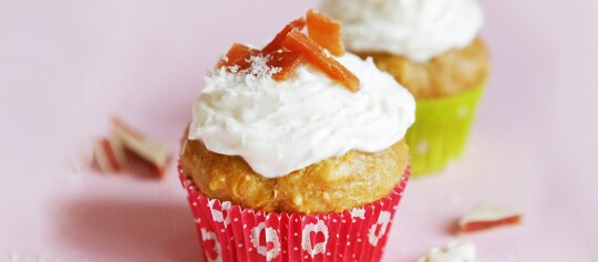Zwei Cupcakes mit weißem Frosting und getrockneten Fruchtstücken, einer in rotem und einer in grünem Papierförmchen, auf rosa Untergrund.