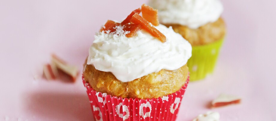 0220_Muffins_DIY_Online_1200x527px4 Zwei Cupcakes mit weißem Frosting und getrockneten Fruchtstücken, einer in rotem und einer in grünem Papierförmchen, auf rosa Untergrund.