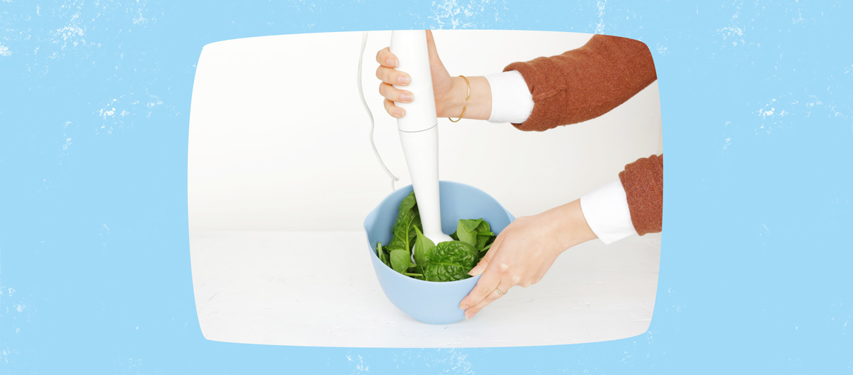 Person verwendet weißen Stabmixer, um frischen Spinat in einer hellblauen Schüssel zu pürieren