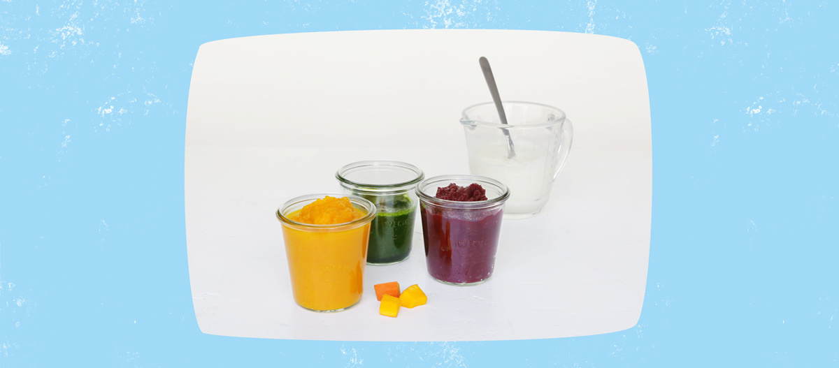 Drei kleine Gläser mit bunten Gemüse- und Obstpürees neben einem Glas mit Joghurt und Löffel auf weißem Hintergrund