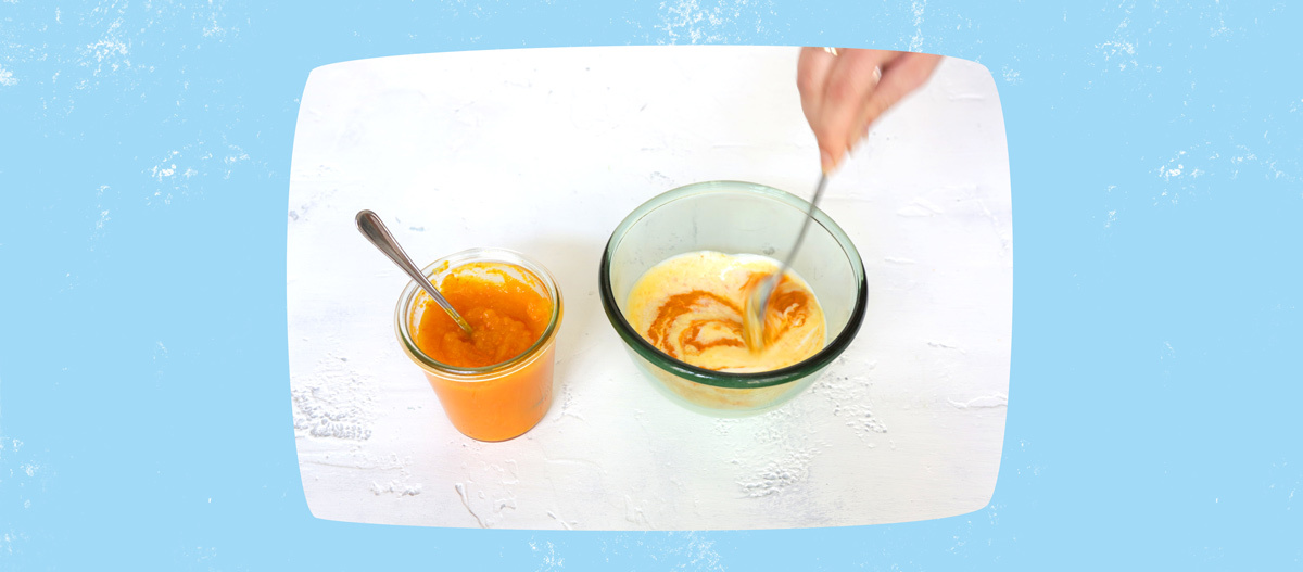 Glasbehälter mit orangem Püree und cremiger Mischung, die mit einem Löffel verrührt wird, auf weißem Untergrund.