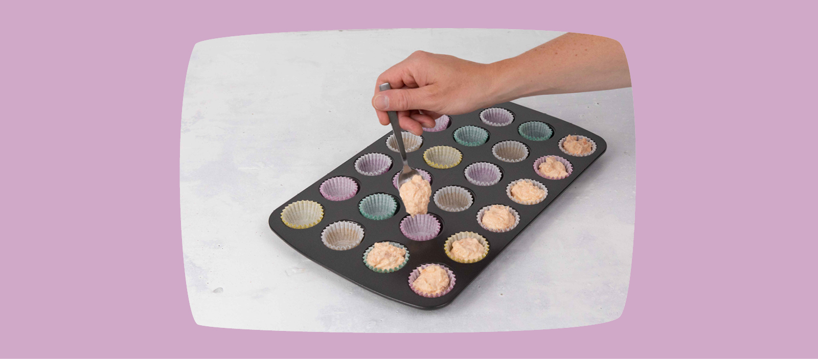 Hand füllt Muffinförmchen mit Teig in einem schwarzen Muffinblech, bunte Papierförmchen, Backvorbereitung