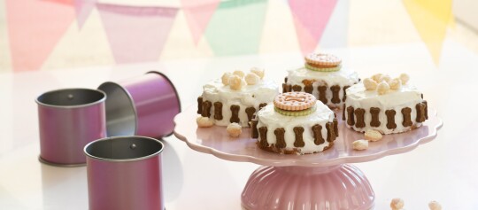 Kleine runde Hundekuchen mit weißem Frosting, verziert mit knochenförmigen Keksen und Hundekeksen auf einem rosa Kuchenständer, neben lila Backformen.