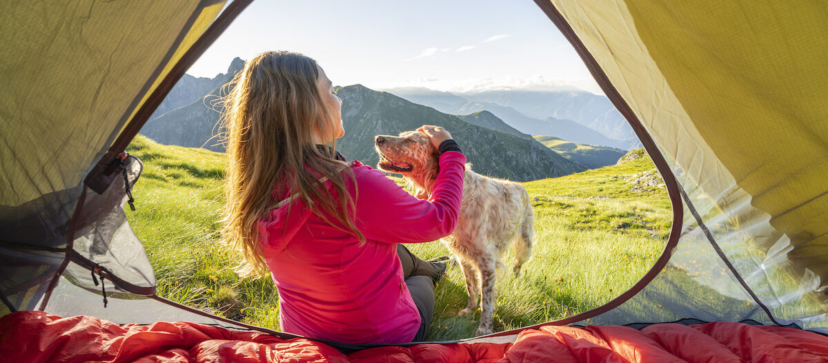 Frau in pinker Jacke streichelt ihren Hund vor einem Zelt auf einer grünen Bergwiese mit Blick auf die Alpen