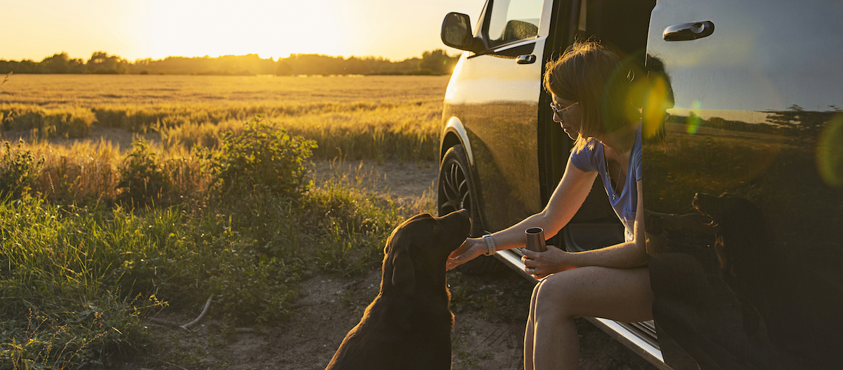 Hund und Halterin machen Pause mit Campingbus am Feldrand Frau sitzt auf der offenen Autotür und streichelt ihren braunen Hund bei Sonnenuntergang auf dem Land