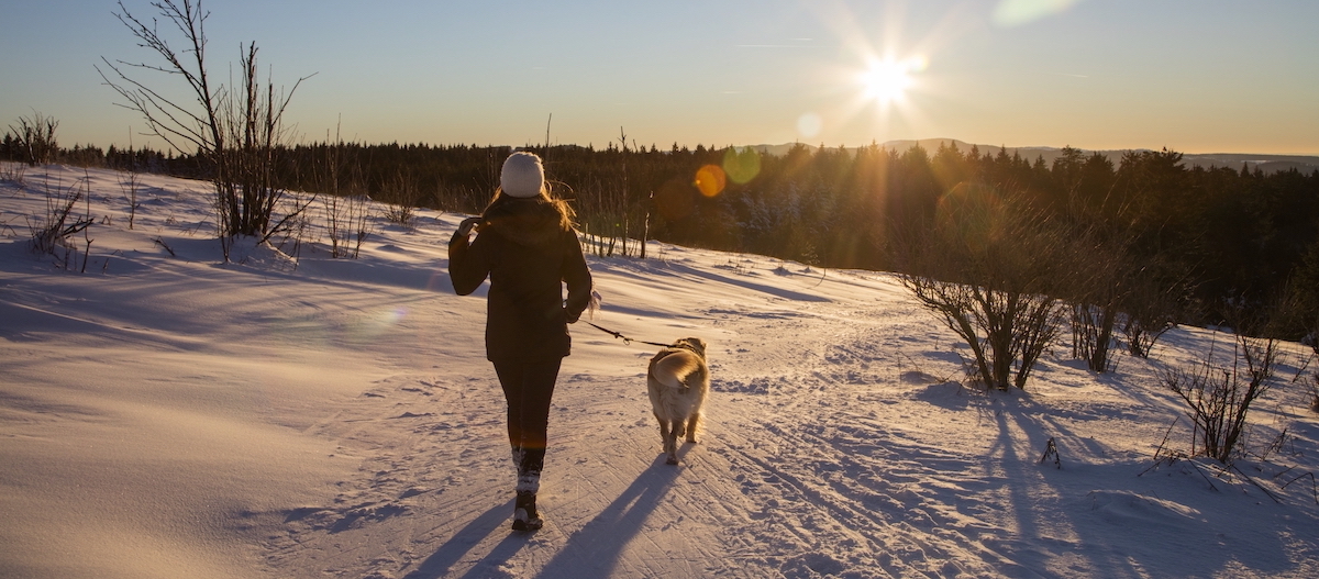 Frau geht mit Hund an der Leine auf verschneitem Weg bei Sonnenuntergang in winterlicher Landschaft