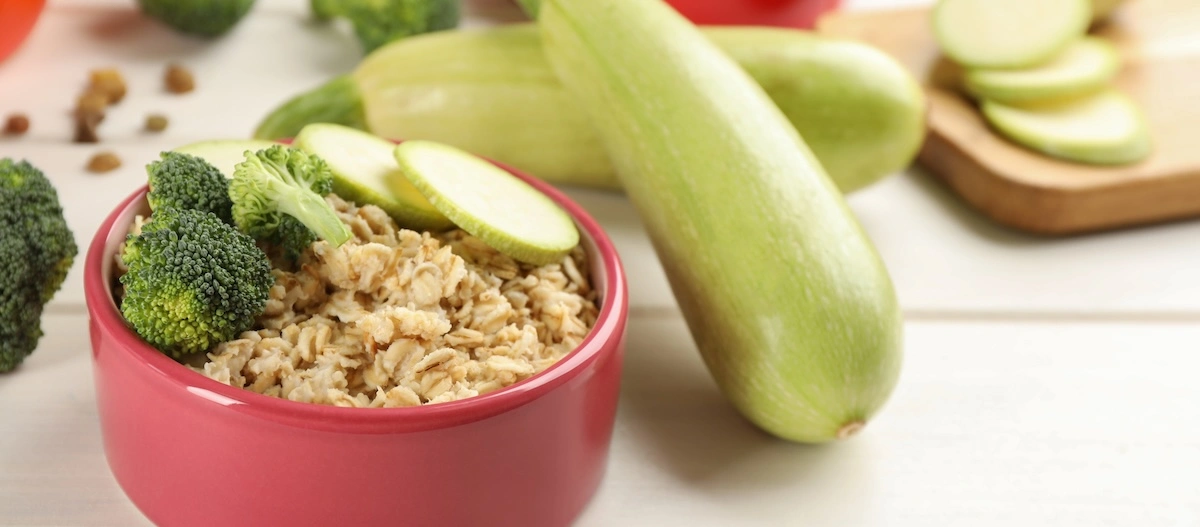 Rote Schüssel mit Haferbrei, garniert mit frischem Brokkoli und Zucchinischeiben, umgeben von ganzen Zucchini und einem Schneidebrett.