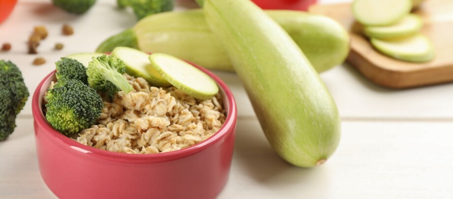 Rote Schüssel mit Haferbrei, garniert mit frischem Brokkoli und Zucchinischeiben, umgeben von ganzen Zucchini und einem Schneidebrett.